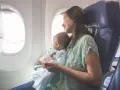 Uśmiechnięta mama z niemowlakiem w ramionach patrzy przez okno samolotu. Ich pierwsza podróż z niemowlakiem pełna jest zachwytu.