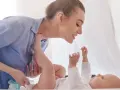 Uśmiechnięta mama pochyla się nad dzieckiem, które leży na plecach. Wykonuje szerokie pieluchowanie, delikatnie unosząc nóżki malucha.
