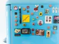 Niebieska lodówka ozdobiona magnesami: Frida Kahlo, renifer, szturmowcy, pocztówka z Madrytu, telewizor, budapeszteński tramwaj i notatnik do druku.