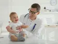 Uśmiechnięty lekarz trzyma na rękach radosne dziecko. Maluch jeszcze nie chodzi, ale jego śmiech rozjaśnia gabinet.