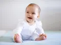 Uśmiechnięte dziecko 8 miesięcy nie siedzi, bawi się stopami na jasnym kocyku.