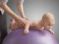 Niemowlę na fioletowej piłce ćwiczy, by zmniejszyć napięcie mięśniowe. Ręce terapeuty delikatnie masują jego plecy.