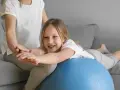 Dziecko z obniżonym napięciem mięśniowym ćwiczy chodzenie na piłce rehabilitacyjnej z pomocą terapeuty.
