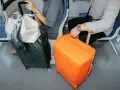 Dwie walizki kabinowe, jedna pomarańczowa, druga czarna, stoją obok siedzących pasażerów w pociągu.