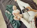 Uśmiechnięte niemowlę na kocyku, mama robi zdjęcie telefonem. To jeden ze sposobów, jak wspierać rozwój niemowlaka – dokumentując jego radość.