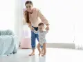 Uśmiechnięta mama pomaga dziecku stawiać pierwsze kroki. Pokazuje, jak trzymać dziecko przy nauce chodzenia, wspierając je za rączki.
