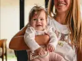 Uśmiechnięta mama trzyma na rękach 5-miesięczne niemowlę. Pokazuje, jak nosić maluszka, który bawi się gryzakiem.