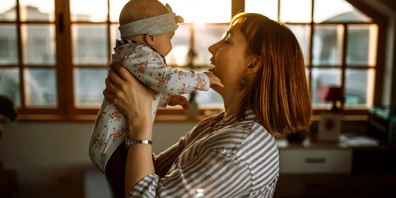 Mama bawi się z dzieckiem, które wykonuje odginanie głowy do tyłu. Ciepłe światło wpada przez okno.