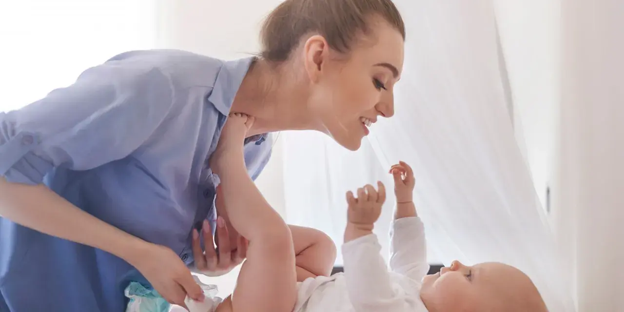 Uśmiechnięta mama pochyla się nad dzieckiem, które leży na plecach. Wykonuje szerokie pieluchowanie, delikatnie unosząc nóżki malucha.
