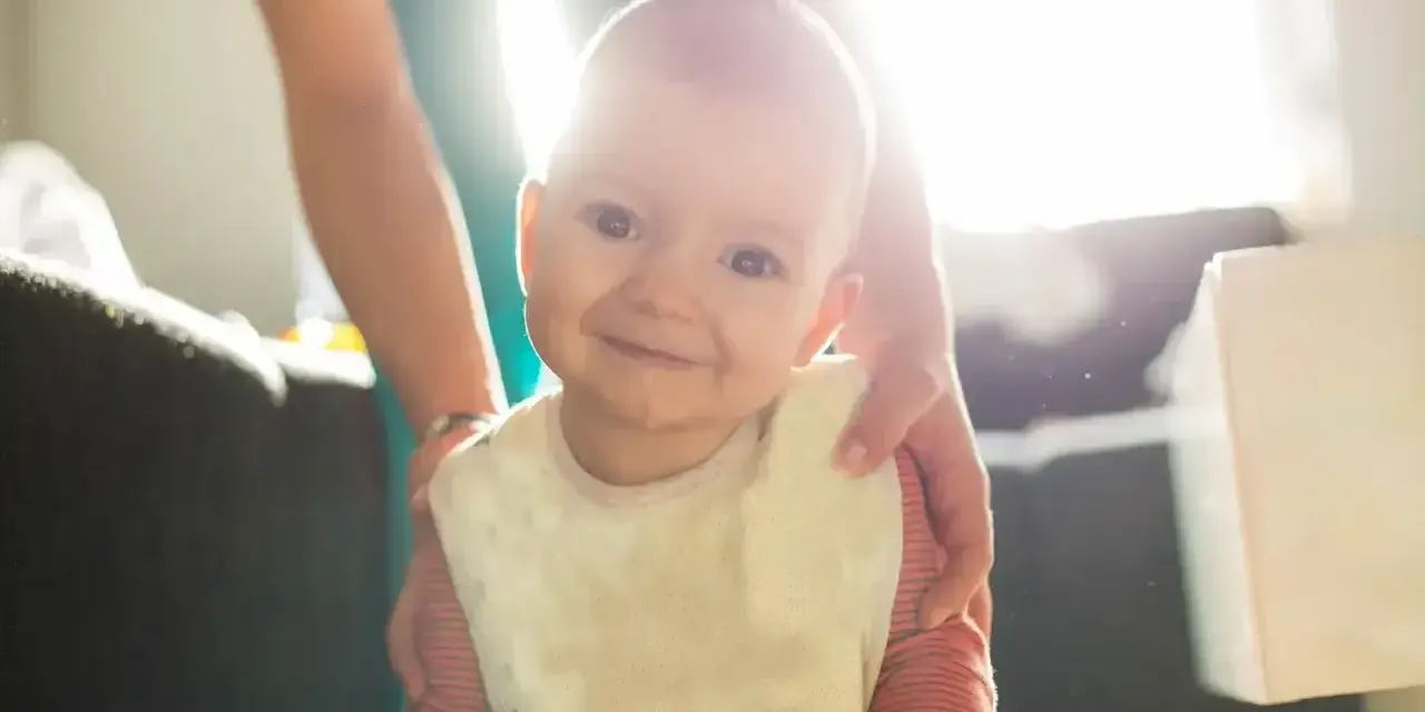 Uśmiechnięte niemowlę wkłada rączki do ust i się ślini, ubrana w śliniaczek.