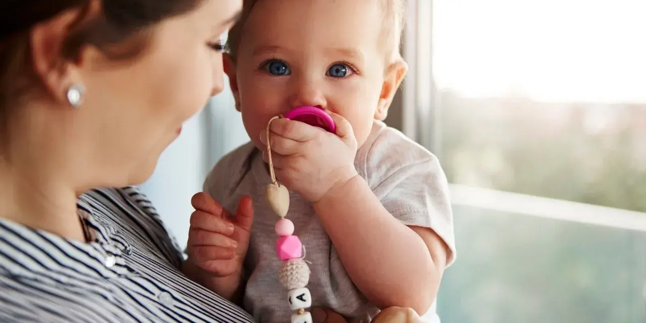 Maluch karmiony piersią z niebieskimi oczami ssie różowy smoczek. Mama trzyma go na rękach, a smoczek ma zawieszkę z imieniem.