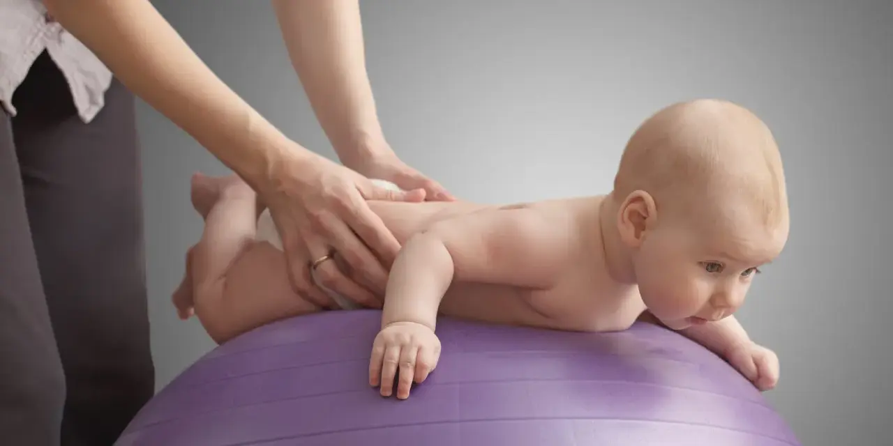 Niemowlę na fioletowej piłce ćwiczy, by zmniejszyć napięcie mięśniowe. Ręce terapeuty delikatnie masują jego plecy.