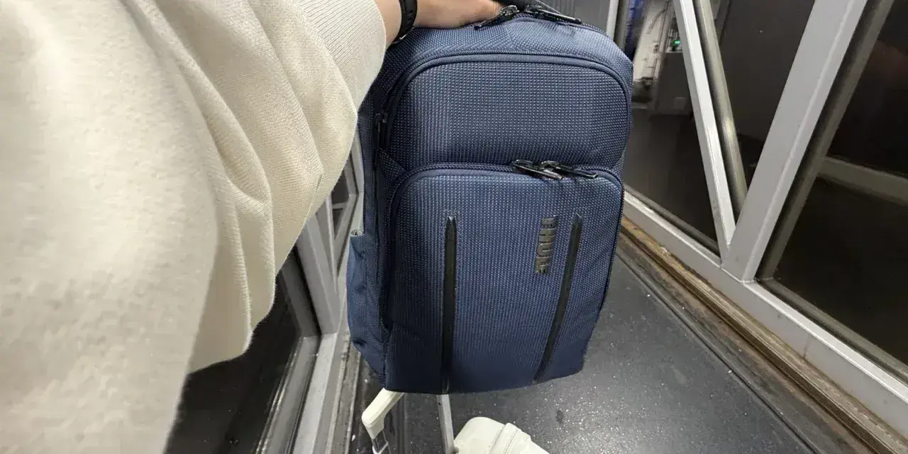 Ręka trzyma granatowy plecak, idealny na podróż z Ryanair. Obok walizka i rękaw lotniska.