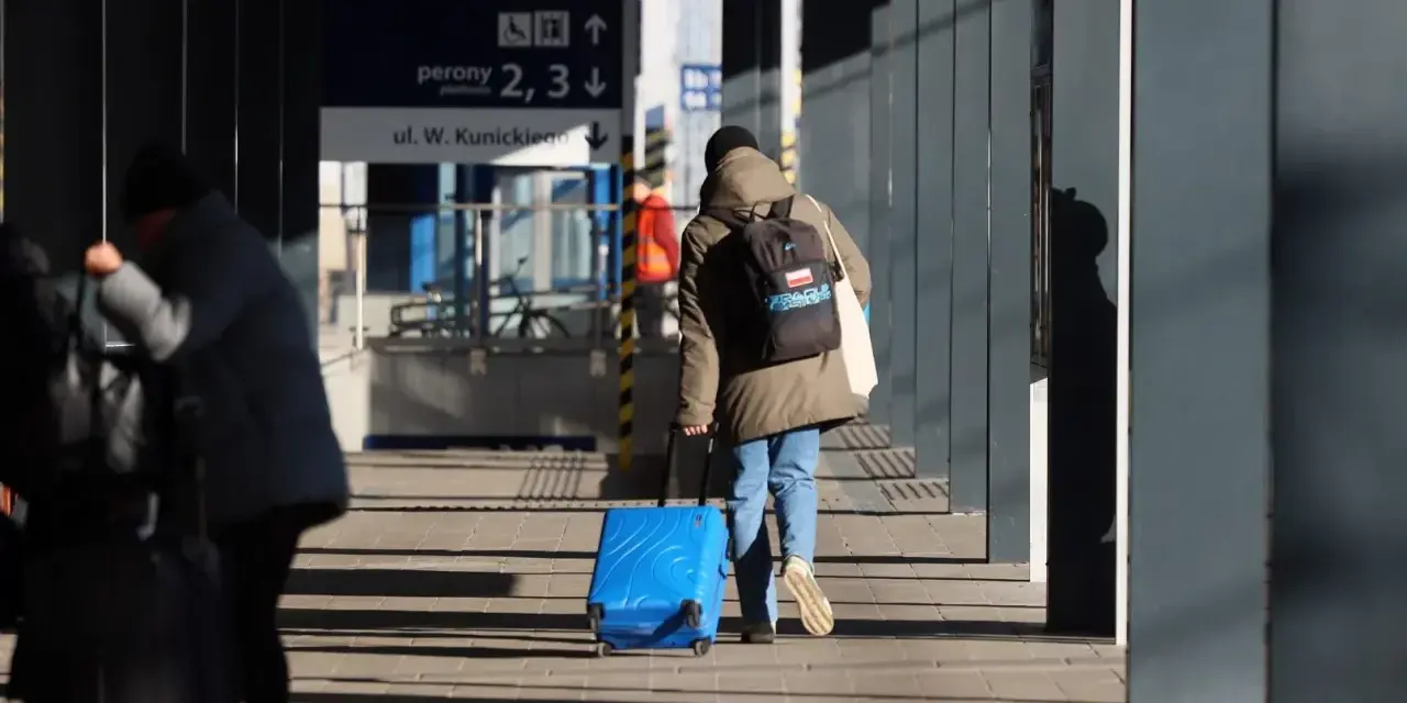 Osoba z plecakiem ciągnie niebieską, intercity dużą walizkę po peronie.