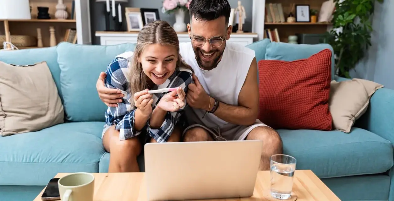 Szczęśliwa para z testem ciążowym na ekranie laptopa. Czy da się zaplanować płeć dziecka? Ich radość sugeruje, że odkryli coś ekscytującego.
