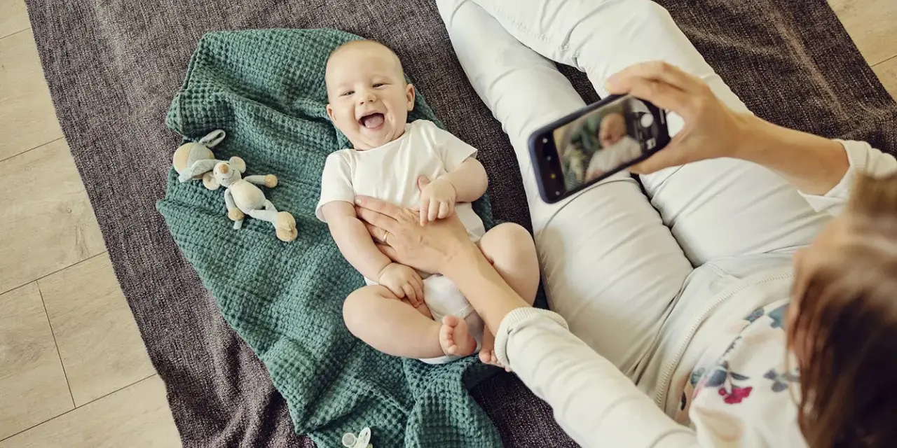 Uśmiechnięte niemowlę na kocyku, mama robi zdjęcie telefonem. To jeden ze sposobów, jak wspierać rozwój niemowlaka – dokumentując jego radość.