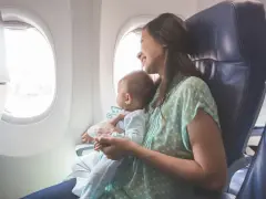Uśmiechnięta mama z niemowlakiem w ramionach patrzy przez okno samolotu. Ich pierwsza podróż z niemowlakiem pełna jest zachwytu.