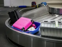 Różowa walizka wypadła z niebieskiego pojemnika, leżąc na taśmie bagażowej lotniska. Obok stoi szara walizka.