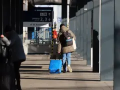 Osoba z plecakiem ciągnie niebieską, intercity dużą walizkę po peronie.