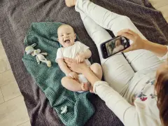 Uśmiechnięte niemowlę na kocyku, mama robi zdjęcie telefonem. To jeden ze sposobów, jak wspierać rozwój niemowlaka – dokumentując jego radość.