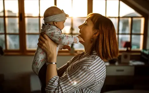 Mama bawi się z dzieckiem, które wykonuje odginanie głowy do tyłu. Ciepłe światło wpada przez okno.