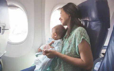 Uśmiechnięta mama z niemowlakiem w ramionach patrzy przez okno samolotu. Ich pierwsza podróż z niemowlakiem pełna jest zachwytu.