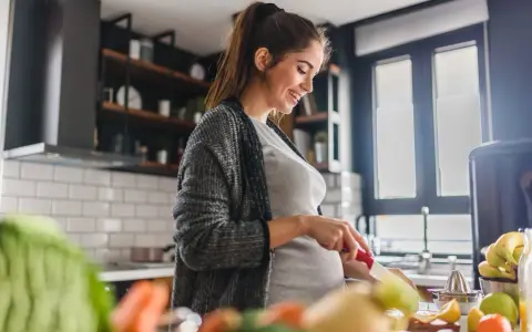 Uśmiechnięta ciężarna kobieta przygotowuje zdrowe jedzenie w kuchni, dbając o to, jak zatrzymać przyrost wagi w ciąży.