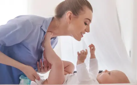 Uśmiechnięta mama pochyla się nad dzieckiem, które leży na plecach. Wykonuje szerokie pieluchowanie, delikatnie unosząc nóżki malucha.