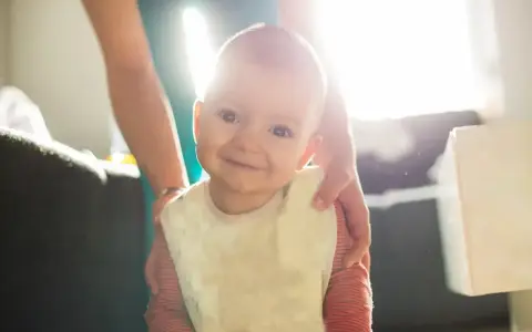 Uśmiechnięte niemowlę wkłada rączki do ust i się ślini, ubrana w śliniaczek.