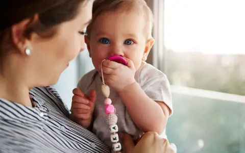 Maluch karmiony piersią z niebieskimi oczami ssie różowy smoczek. Mama trzyma go na rękach, a smoczek ma zawieszkę z imieniem.