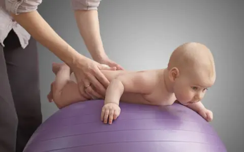 Niemowlę na fioletowej piłce ćwiczy, by zmniejszyć napięcie mięśniowe. Ręce terapeuty delikatnie masują jego plecy.