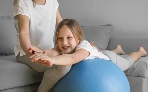 Dziecko z obniżonym napięciem mięśniowym ćwiczy chodzenie na piłce rehabilitacyjnej z pomocą terapeuty.