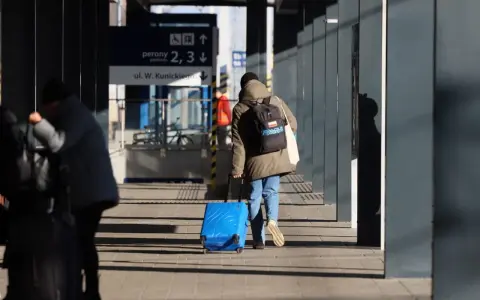 Osoba z plecakiem ciągnie niebieską, intercity dużą walizkę po peronie.