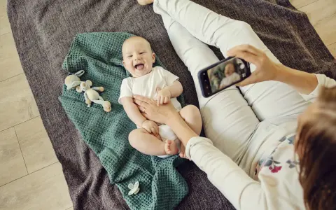 Uśmiechnięte niemowlę na kocyku, mama robi zdjęcie telefonem. To jeden ze sposobów, jak wspierać rozwój niemowlaka – dokumentując jego radość.