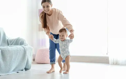 Uśmiechnięta mama pomaga dziecku stawiać pierwsze kroki. Pokazuje, jak trzymać dziecko przy nauce chodzenia, wspierając je za rączki.