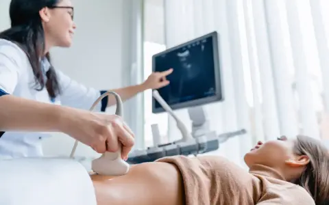 Lekarka wykonuje badania prenatalne 1 trymestr, przykładając głowicę USG do brzucha pacjentki. Na ekranie widoczny obraz.