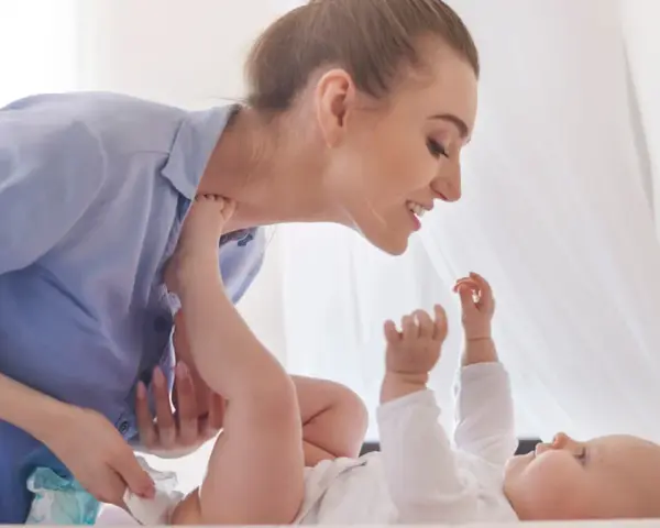 Uśmiechnięta mama pochyla się nad dzieckiem, które leży na plecach. Wykonuje szerokie pieluchowanie, delikatnie unosząc nóżki malucha.