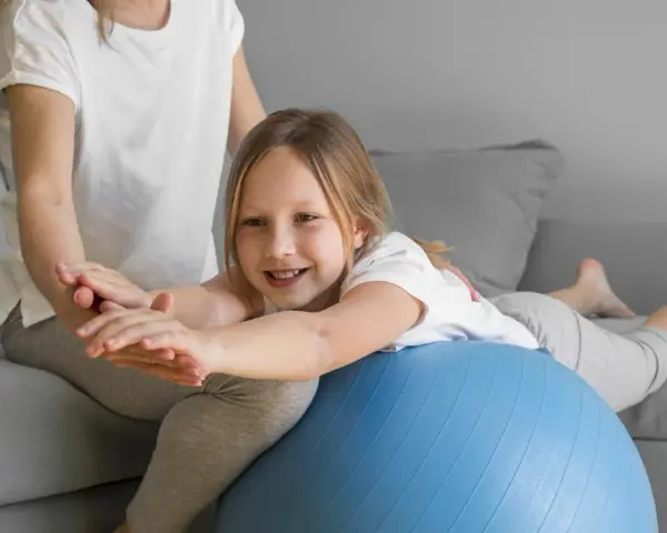 Dziecko z obniżonym napięciem mięśniowym ćwiczy chodzenie na piłce rehabilitacyjnej z pomocą terapeuty.