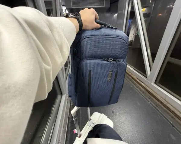 Ręka trzyma granatowy plecak, idealny na podróż z Ryanair. Obok walizka i rękaw lotniska.