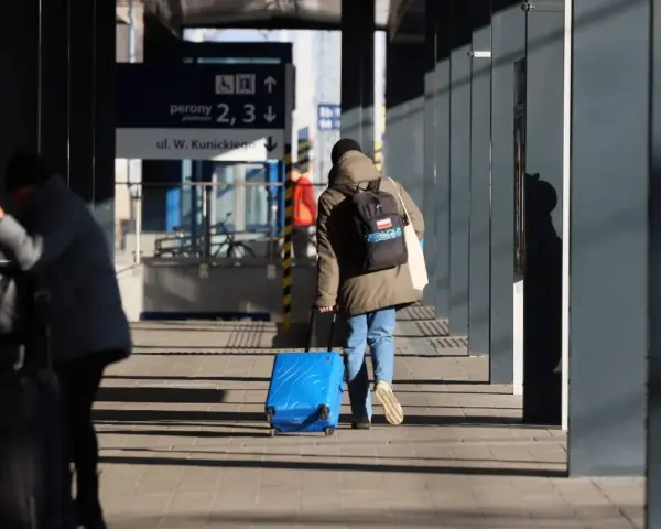 Osoba z plecakiem ciągnie niebieską, intercity dużą walizkę po peronie.