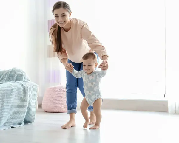 Uśmiechnięta mama pomaga dziecku stawiać pierwsze kroki. Pokazuje, jak trzymać dziecko przy nauce chodzenia, wspierając je za rączki.