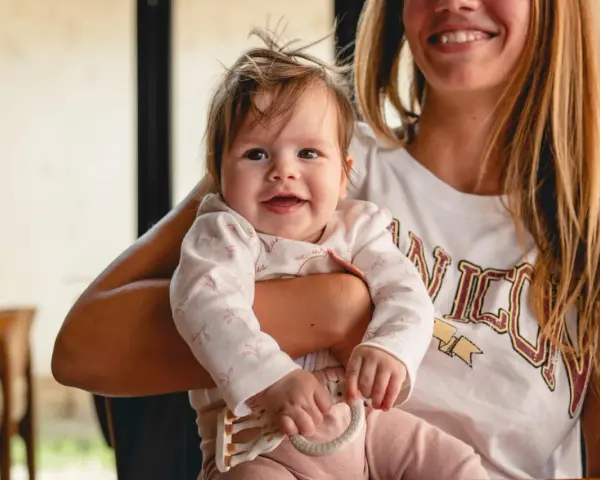 Uśmiechnięta mama trzyma na rękach 5-miesięczne niemowlę. Pokazuje, jak nosić maluszka, który bawi się gryzakiem.