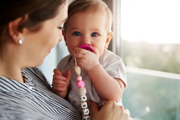 Maluch karmiony piersią z niebieskimi oczami ssie różowy smoczek. Mama trzyma go na rękach, a smoczek ma zawieszkę z imieniem.