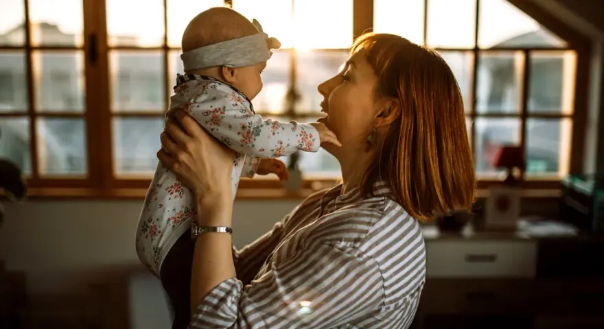 Mama bawi się z dzieckiem, które wykonuje odginanie głowy do tyłu. Ciepłe światło wpada przez okno.
