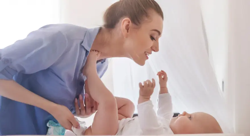Uśmiechnięta mama pochyla się nad dzieckiem, które leży na plecach. Wykonuje szerokie pieluchowanie, delikatnie unosząc nóżki malucha.