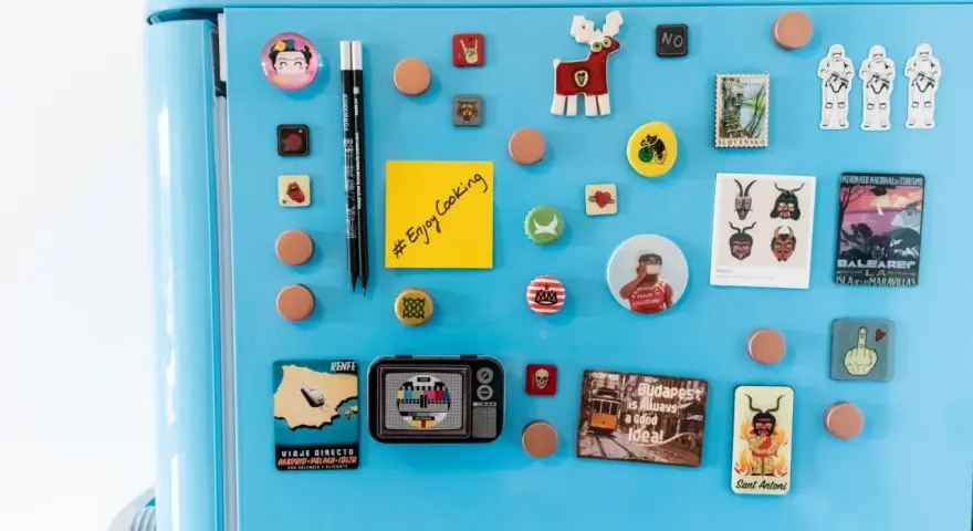 Niebieska lodówka ozdobiona magnesami: Frida Kahlo, renifer, szturmowcy, pocztówka z Madrytu, telewizor, budapeszteński tramwaj i notatnik do druku.