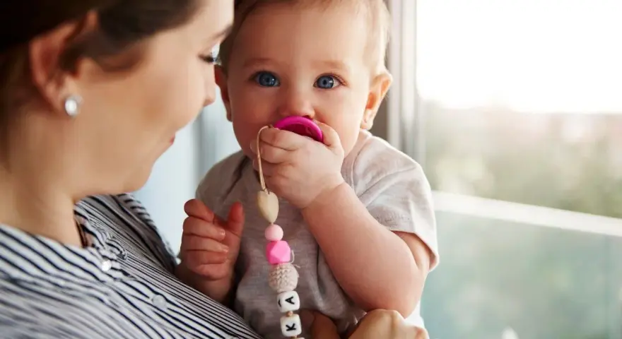 Maluch karmiony piersią z niebieskimi oczami ssie różowy smoczek. Mama trzyma go na rękach, a smoczek ma zawieszkę z imieniem.