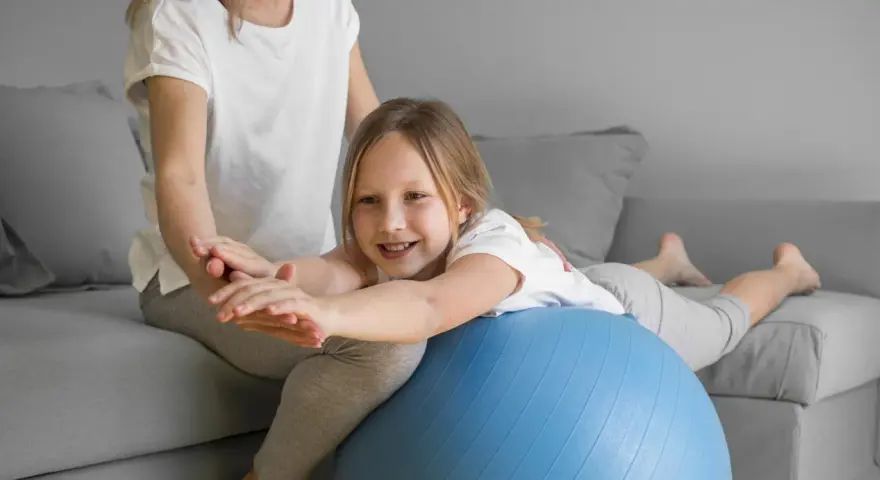 Dziecko z obniżonym napięciem mięśniowym ćwiczy chodzenie na piłce rehabilitacyjnej z pomocą terapeuty.