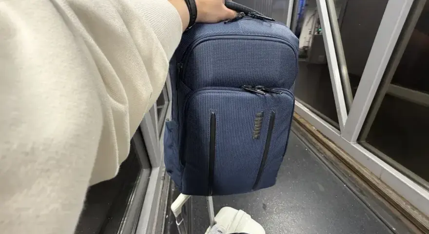 Ręka trzyma granatowy plecak, idealny na podróż z Ryanair. Obok walizka i rękaw lotniska.