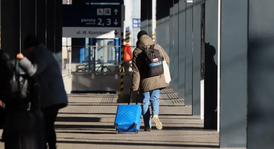 Osoba z plecakiem ciągnie niebieską, intercity dużą walizkę po peronie.